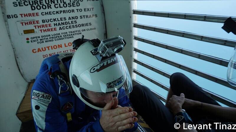 Brian Germain in a helmet at the open door of an aircraft, sky visible behind him